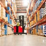 Worker in forklift-truck loading packed goods in huge distribution warehouse with high shelves.